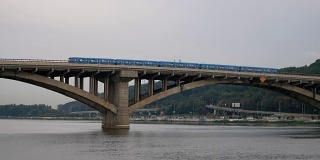火车在天空和森林的映衬下驶过大桥。城市地铁就在外面。公共交通在移动。汽车在河上的桥上行驶。