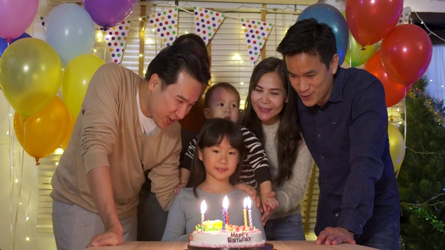 一个可爱的小女孩坐在桌子旁，吹着生日蛋糕上的蜡烛，而她的家人站在后面为她唱歌。她给了一个大大的微笑，她的家人为她鼓掌。在缓慢的运动。