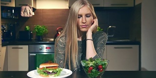 年轻女子在汉堡包和沙拉之间做出选择。漂亮的年轻女士坐在时髦的厨房里看着美味的汉堡和健康的蔬菜沙拉
