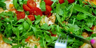 特写，大而多汁的热披萨，配上绿色蔬菜，芝麻菜和樱桃番茄。女性用刀叉切披萨