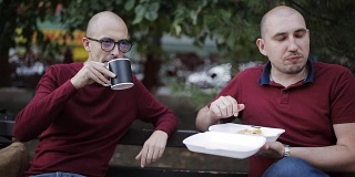 两个男人坐在公园的长椅上边吃三明治边聊天