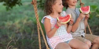 快乐的女孩和男孩坐在秋千上吃着西瓜，夏天的照片。孩子们拿着西瓜