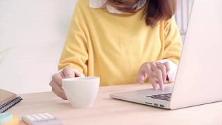 美丽的年轻微笑的亚洲女人工作在笔记本电脑和喝咖啡在家里的客厅。亚洲商业女性工作文件财务和计算器在她的家庭办公室。享受在家的时光。视频素材模板下载