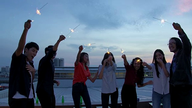 一群快乐的年轻朋友享受和玩火花在屋顶聚会傍晚日落。节日庆祝节日派对。青少年的生活方式。缓慢的运动。