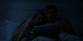 女士和男士坐在黑房间的床上，用手提电脑看恐怖电影