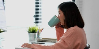 年轻的亚洲女性打字键盘和喝咖啡在家庭办公室与快乐的情绪，在家工作，小企业，在线商务，在线教育，办公室休闲的生活方式概念