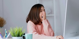 年轻的亚洲女人打哈欠，疲惫的工作与电脑在她的家庭办公室，在家里工作，人们在办公室的生活方式