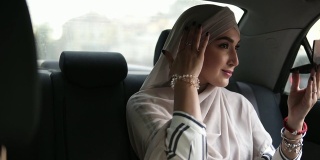 一个年轻的穆斯林妇女在米色头巾的肖像，坐在车里，看着一个小化妆镜，检查她的化妆