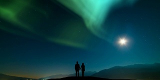 这对夫妇站在山上，映衬着北极光的天空。时间流逝