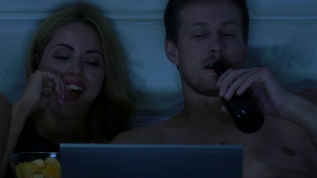 年轻夫妇在床上用笔记本电脑看连续剧吃零食喝啤酒