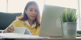 美丽的年轻微笑的亚洲女人工作在笔记本电脑和喝咖啡在家里的客厅。亚洲商业女性工作文件财务和计算器在她的家庭办公室。享受在家的时光。