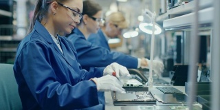 女电子工厂的工人在蓝色的工作外套和保护眼镜组装笔记本电脑的主板与螺丝刀。拥有多名员工的高科技工厂设施。