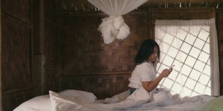 年轻女子坐在卧室白色的床上用智能手机，享受微风吹拂她的头发。