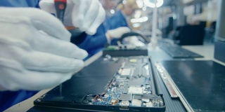一个穿着蓝色工作服的电子工厂女工用螺丝刀组装笔记本电脑的主板的特写。拥有多名员工的高科技工厂设施。
