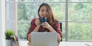 年轻的亚洲女性工作与笔记本电脑和喝咖啡在家办公与快乐的情绪，在家里工作，小企业，在线商务，在线教育，办公室休闲的生活方式概念