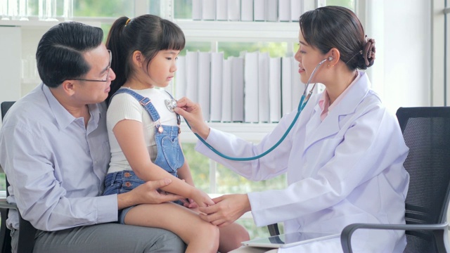 女儿科医生用听诊器检查可爱的小女孩。孩子开心地笑着指着医生。父亲抱着她的孩子。中国和香港的医疗系统