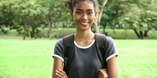 年轻迷人的非洲裔美国混合妇女微笑着双臂交叉与学校背包袋在公园户外