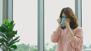 年轻美丽的亚洲女人喝咖啡，透过窗户与城市的看法与幸福，人们的生活方式视频素材模板下载