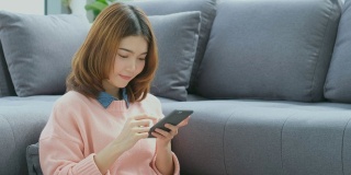 年轻美丽的亚洲女人使用智能手机与幸福坐在沙发上，人们在电话，生活方式