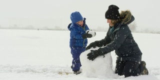 3岁的白人男孩和他的白人母亲在她30多岁(都穿着冬天的衣服)一起堆雪人在一个下雪，阴天