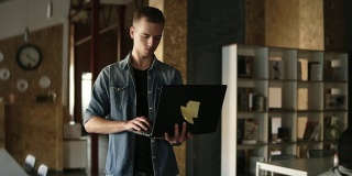 有吸引力的集中的年轻商人正走在他的手中黑色笔记本电脑和打字。白种人在公共场所工作，内部是砖墙。缓慢的运动。前视图
