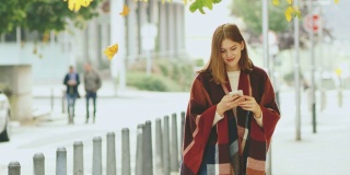 一名年轻女子穿着格子羊绒披肩，一边走在秋天的人行道上，一边用智能手机发短信
