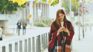 一名年轻女子穿着格子羊绒披肩，一边走在秋天的人行道上，一边用智能手机发短信视频素材模板下载