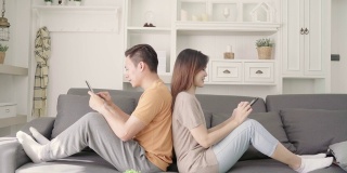 亚洲情侣在家中客厅用智能手机查看社交媒体，甜蜜的情侣在家中放松时躺在沙发上享受爱情时刻。生活方式夫妇在家里放松的概念。