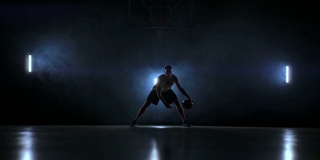 一名男子在黑暗的篮球场上拿着篮球，背景是烟雾中的篮球圈，在三盏背光灯的照耀下，他展示着运球技巧