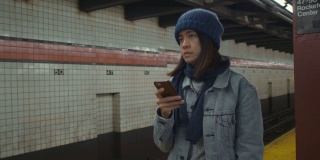 年轻的亚洲女子在地铁站台上用手机等待和发短信。