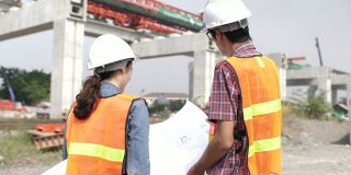 两个年轻的工程师在工地一起聊天，背景是大吊车，他们穿着安全设备