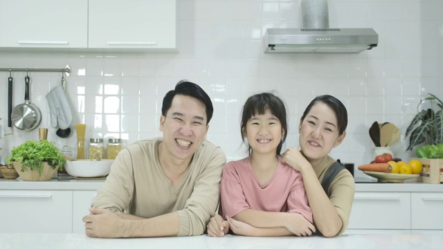 幸福的亚洲家庭，母亲，父亲和女儿坐在厨房和微笑在镜头前。肖像拍摄。在室内