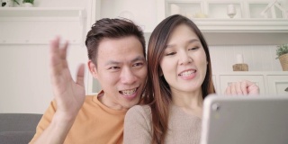 亚洲情侣在家中客厅用平板电脑与朋友视频通话，甜蜜情侣在家中放松时躺在沙发上享受爱情时刻。生活方式夫妇在家里放松的概念。