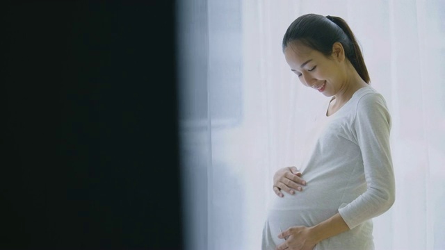 美丽的亚洲孕妇透过窗户看，并在客厅揉胃。