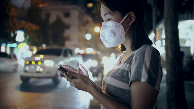 女人戴着口罩，在晚上使用智能手机来保护自己免受空气污染