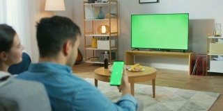 一对幸福的夫妇坐在客厅里看绿色屏幕电视，在沙发上放松。Guy还使用绿色模拟屏幕智能手机。