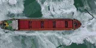 在极端冬季条件下在冰冻海面上航行的货船航拍。在破冰船造成的狭窄航道中航行。北方寒冷冬季的水运