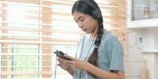 年轻美丽的亚洲女性戴着耳机和智能手机站在厨房，人和技术，Z一代的生活方式