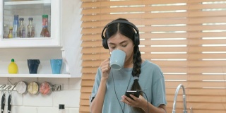 年轻美丽的亚洲女人戴着耳机，用智能手机在厨房喝咖啡，人和科技，Z世代的生活方式