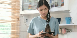 年轻美丽的亚洲女人站在厨房里使用平板电脑和耳机，人们和科技，Z一代的生活方式