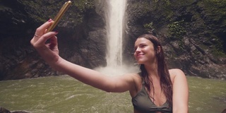 年轻女子旅游博主做自拍手机上的山瀑布背景。微笑的女人拍摄的手机自拍在飞溅的瀑布流动在雨林。