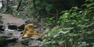 穿着黄色雨衣的亚洲女孩坐在瀑布，张开双臂。