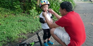 爸爸给2岁的亚洲小孩戴上护膝，爸爸和儿子在公园里玩平衡自行车(跑步自行车)