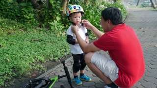 爸爸给2岁的亚洲小孩戴上护膝，爸爸和儿子在公园里玩平衡自行车(跑步自行车)视频素材模板下载