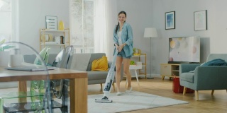 年轻漂亮的女人在一个明亮舒适的房间在家里跳舞和吸尘机地毯。她用的是现代无绳吸尘器。她是快乐和快乐的。