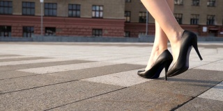 在阳光明媚的日子里，穿着黑色高跟鞋的苗条女性的腿走在城市广场上。年轻女商人的脚穿着高跟鞋走在城市的街道上。低角度视角慢动作特写