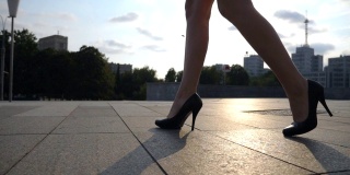 在日落时分，穿着黑色高跟鞋走在城市广场上的苗条女性腿。年轻女商人的脚穿着高跟鞋走在城市的街道上。低角度视角慢动作特写