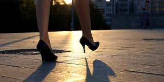 跟随女性的腿高跟鞋走在城市街道在日落时间。穿着高跟鞋的女商人的脚在城市里走动。女孩走着去上班。慢动作关闭侧视图
