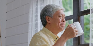 年长的亚洲男人站在窗边喝咖啡，放松情绪，退休生活