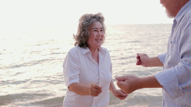 亚洲高年级情侣在沙滩上跳舞一起笑。有生活方式、情感和退休观念的人。慢动作镜头。